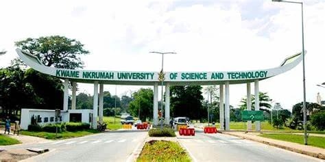 A wide shot of the iconic main entrance gate of the Kwame Nkrumah University of Science and Technology (KNUST) in Kumasi. The white curved structure features the university's name in green lettering, overlooking a road with light traffic and lush greenery.