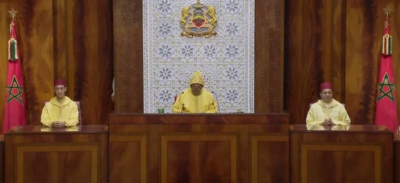 King Mohammed VI of Morocco delivers a speech from a central podium, flanked by Crown Prince Moulay El Hassan and Prince Moulay Rachid. They are dressed in traditional yellow robes and red fezzes within a formal setting featuring the Moroccan royal coat of arms and national flags.