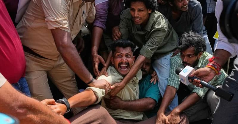 A group of young protesters are involved in a physical struggle with authorities, with a man in the center shouting intensely while being restrained. Several other individuals are seen in the chaotic, crowded scene, including a police officer in a khaki uniform and a reporter holding a microphone towards the action.