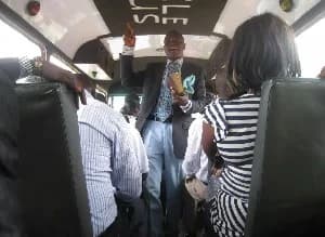 A man dressed in a suit and tie stands in the aisle of a public transport bus, gesturing as he appears to be preaching to the seated passengers. The perspective is from the back of the vehicle, showing passengers in their seats facing the speaker.