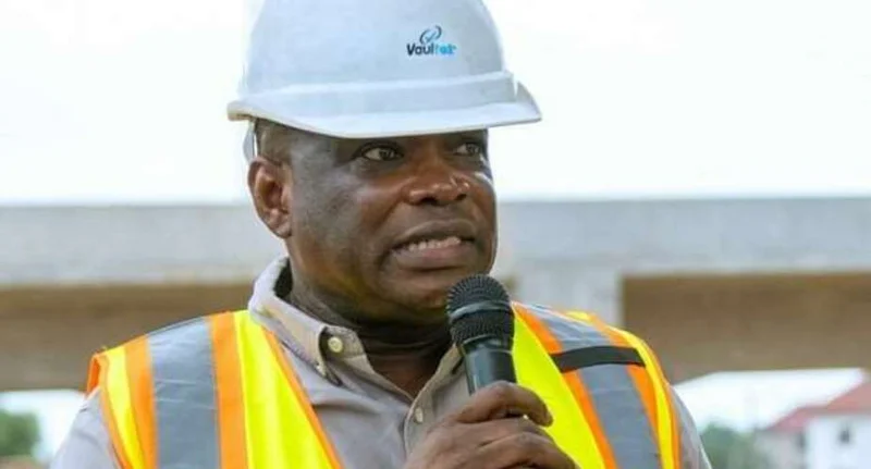 A close-up photograph of a man wearing a white safety hard hat and a high-visibility yellow and orange reflective vest speaking into a microphone. He appears to be at an outdoor construction site with a concrete bridge or infrastructure project visible in the blurred background.