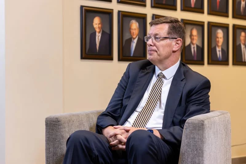 A man in a navy suit and striped tie sits in a grey armchair, looking off-camera during what appears to be an interview. Behind him, a beige wall features several framed portraits of men in formal attire, which are characteristic of leadership galleries in institutional settings.
