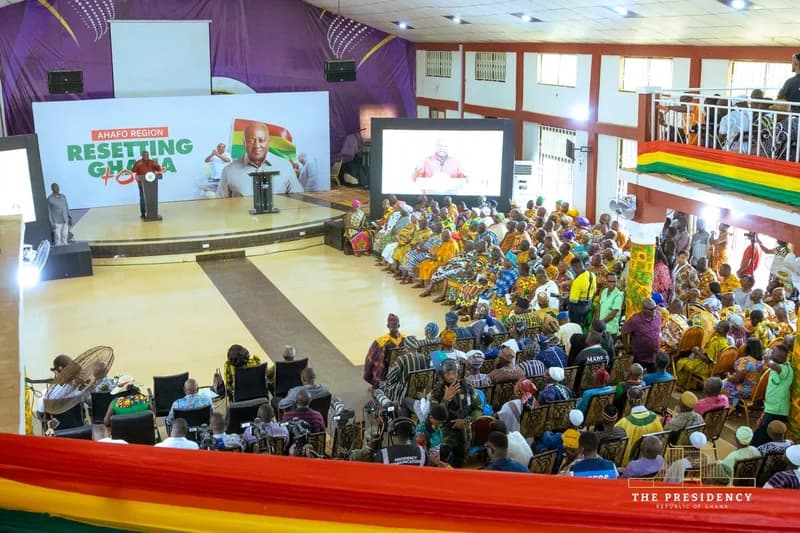 President John Dramani Mahama addresses a large audience during his 'Resetting Ghana Tour' in the Ahafo Region. The event features a diverse crowd, including traditional rulers in colorful Kente cloth, gathered in a large hall with official branding and digital screens displaying the President's speech.
