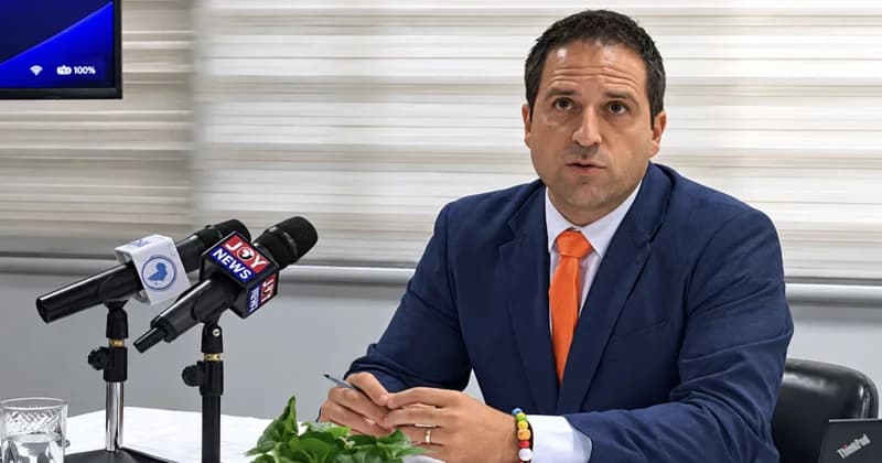 A man in a navy blue suit and bright orange tie, identified as UN Special Rapporteur Balakrishnan Rajagopal, speaks at a press conference in Ghana. He is seated behind microphones, including one from Joy News, while holding a pen and addressing housing affordability issues.