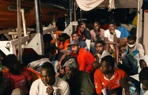 A group of African men are seen huddled together in a cramped, poorly lit environment, likely the interior or deck of a boat. They appear weary and are dressed in casual clothing, including t-shirts, hoodies, and hats, reflecting the difficult conditions of irregular migration.