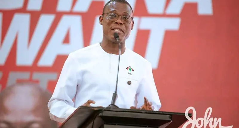 Fifi Fiavi Kwetey, the General Secretary of the National Democratic Congress (NDC), is seen delivering a speech from a podium. He is dressed in a white shirt with an NDC emblem, positioned in front of a red political backdrop featuring campaign text and the name 'John'.