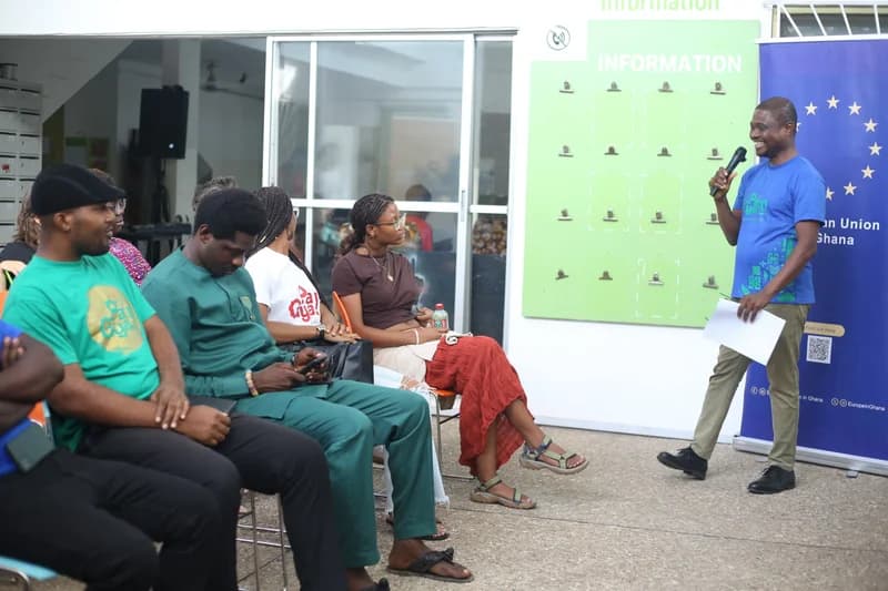 A speaker in a blue t-shirt addresses a seated audience during a session at the Pa Gya! Festival in Ghana. The attendees are listening attentively in an outdoor setting featuring European Union Ghana promotional materials and a green information board.