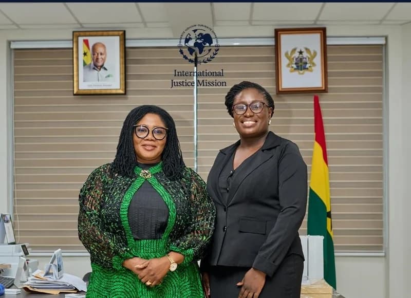 Two women stand smiling in a professional office setting in Ghana. The background features the Ghanaian flag, the national Coat of Arms, a portrait of President John Mahama, and the logo for International Justice Mission (IJM).