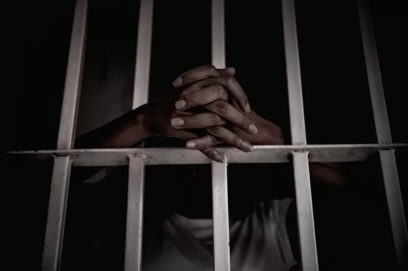 A close-up photograph showing a person's hands clasped together behind white metal prison bars. The background is dark, emphasizing the theme of incarceration and the legal consequences of a crime.