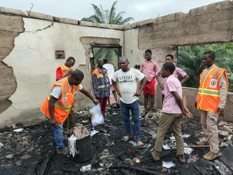 NADMO officials and local residents assess the extensive damage inside a school building following a fire disaster. The floor is covered in charred debris and ash, with officials in orange high-visibility vests inspecting burnt documents while students observe the scene.