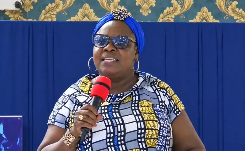 A woman wearing a vibrant blue headwrap and sunglasses speaks into a red microphone. She is dressed in a blue and yellow patterned top and stands before a background consisting of a blue curtain and gold floral wallpaper.