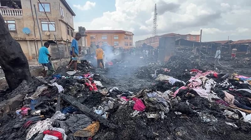 The image depicts the devastating aftermath of a fire in a commercial area, likely in Kumasi, Ghana. Piles of charred debris and partially burnt clothing cover the ground while smoke still rises from the ruins. Residents and onlookers are seen walking through the site, surveying the extensive damage to the market stalls and nearby buildings.