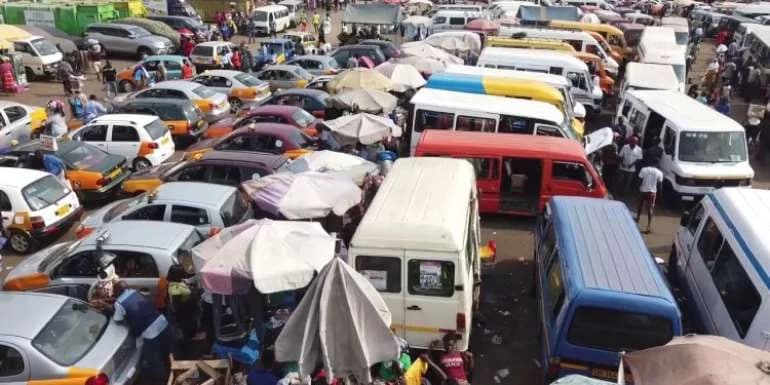 A crowded transportation hub in Ghana, likely a lorry park, filled with numerous trotros and taxis parked closely together. Market umbrellas are scattered among the vehicles, and people are seen moving through the congested area, illustrating urban transport infrastructure challenges.