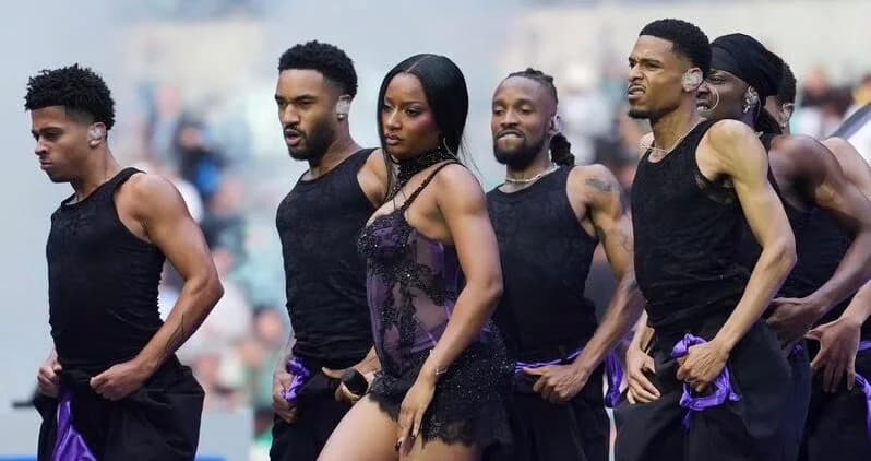 Nigerian singer Ayra Starr performs on stage during the NFL London halftime show, surrounded by a troupe of male backup dancers. The performers are wearing coordinated black and purple outfits while delivering a high-energy Afrobeats performance in a stadium setting.