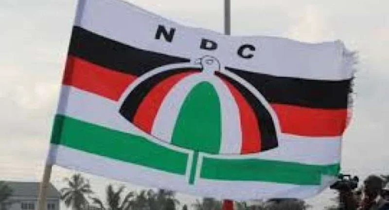 The official flag of the National Democratic Congress (NDC) is seen waving outdoors, featuring its signature umbrella logo and horizontal stripes of black, red, white, and green. In the background, there is a cloudy sky, trees, and glimpses of people with cameras, suggesting a political event or gathering.