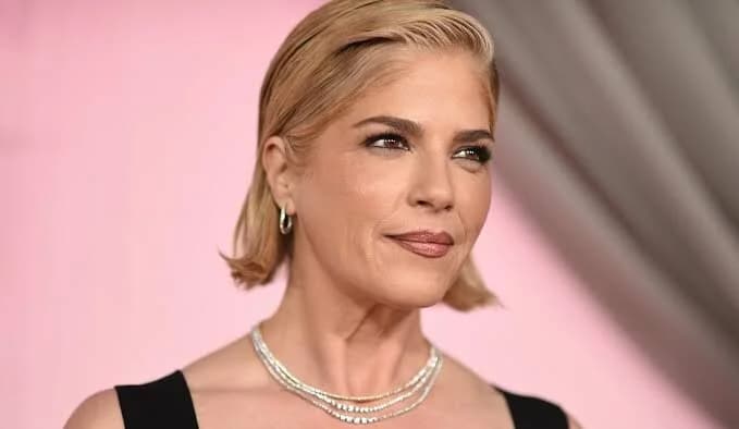 A close-up portrait of American actress Selma Blair looking slightly to the right of the frame. She features short blonde hair and is wearing a layered silver necklace against a soft pink and grey blurred background.