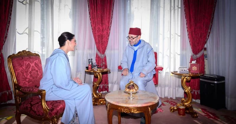 King Mohammed VI of Morocco sits in a formal meeting room, engaging in conversation with a woman. Both individuals are dressed in traditional light blue djellabas, with the King wearing a signature red fez, set against a backdrop of ornate red and gold decor.