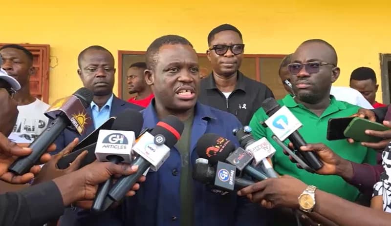 Sam Nartey George, a Ghanaian Member of Parliament, addresses the media during a press conference. He is surrounded by journalists holding multiple microphones and recording devices against a bright yellow background wall.