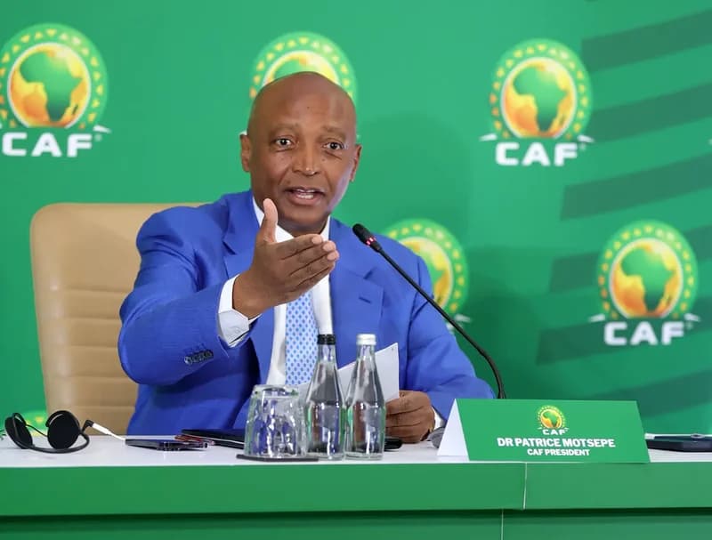 CAF President Dr. Patrice Motsepe is captured speaking at a press conference, gesturing with his hand while seated at a podium. He is wearing a blue suit and white shirt, set against a vibrant green backdrop adorned with multiple CAF logos.