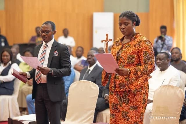 A photograph capturing a formal swearing-in ceremony in Ghana. A man in a suit and a woman in a traditional orange patterned dress are seen standing and reading from documents, with the woman holding a crucifix and the man holding a Bible.