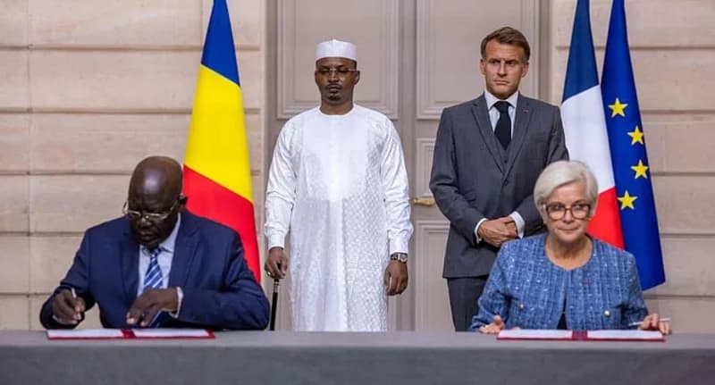French President Emmanuel Macron and Chadian President Mahamat Idriss Déby Itno stand behind two officials signing documents during a formal ceremony. The national flags of Chad and France are positioned behind the leaders, symbolizing the diplomatic agreement between the two nations.