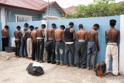 A group of approximately twelve young men are seen lined up against a light blue wall during what appears to be a law enforcement operation. Most of the individuals are shirtless and have their heads bowed toward the wall in an outdoor courtyard setting.