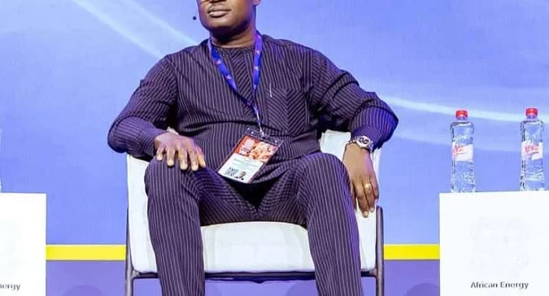 A man, likely Ghana's Energy Minister, sits on a white chair during an 'African Energy' event or conference. He is dressed in a dark pinstriped traditional attire with a conference lanyard around his neck. The setting includes a purple background and water bottles on a side table.