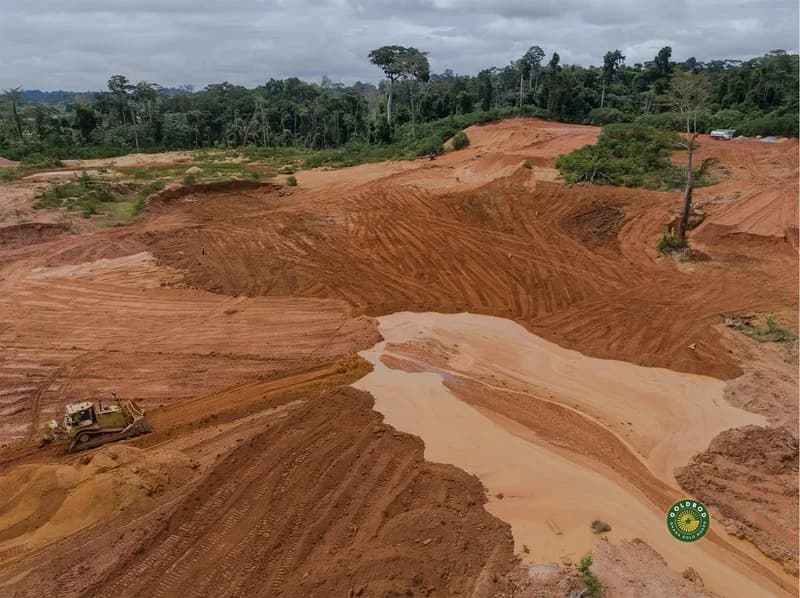 A wide-angle photograph showing a massive area of excavated reddish-brown earth and a pool of muddy water, likely a galamsey site undergoing reclamation. A bulldozer is seen working on the left side of the frame against a backdrop of dense tropical forest.