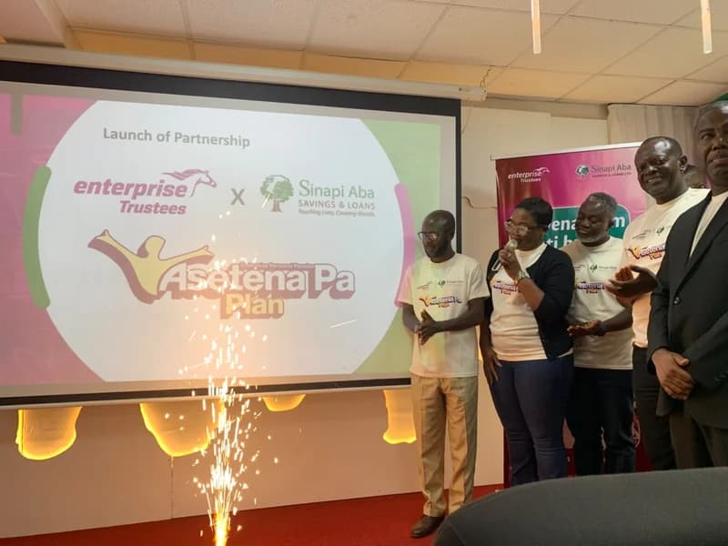 A group of officials stand next to a projector screen during the launch of the Asetena Pa Plan partnership between Sinapi Aba Savings & Loans and Enterprise Trustees. A small pyrotechnic sparkler is active in the foreground as a woman speaks into a microphone while others in branded t-shirts look on.