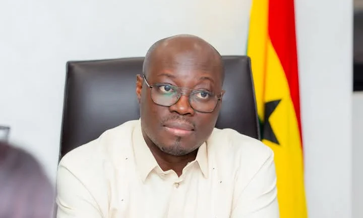 A portrait of Ghanaian politician Dr. Cassiel Ato Forson seated in an official setting. He is wearing glasses and a cream-colored shirt, with the Ghanaian flag visible in the background.