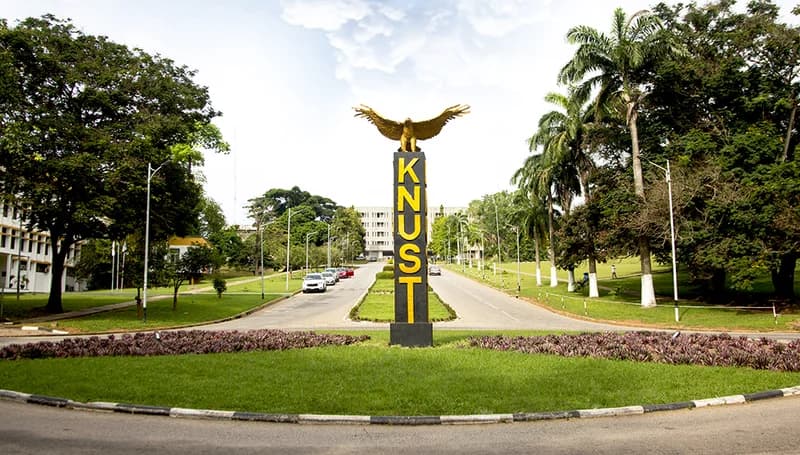 A wide-angle photograph of the iconic entrance to the Kwame Nkrumah University of Science and Technology (KNUST) in Kumasi, Ghana. The central monument features a tall black pillar with vertical yellow 'KNUST' lettering, topped with a golden eagle statue, surrounded by lush green lawns and tropical palm trees.