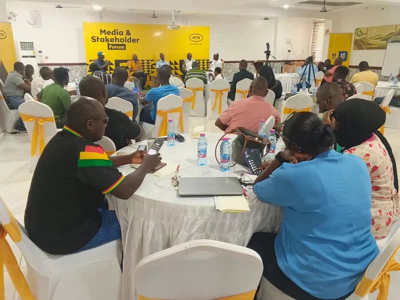 A large group of people are gathered at a Media and Stakeholder Forum hosted by MTN Ghana. Participants are seated at white-clothed tables decorated with yellow bows, facing a panel of speakers in front of a bright yellow MTN branded backdrop.