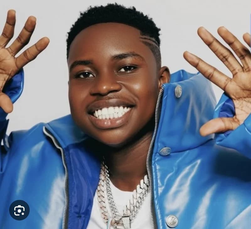 A vibrant close-up portrait of the Nigerian content creator Peller, who is seen smiling broadly and gesturing with both hands raised. He is dressed in a shiny bright blue puffer jacket layered over a white t-shirt and accessorized with chunky silver chains.