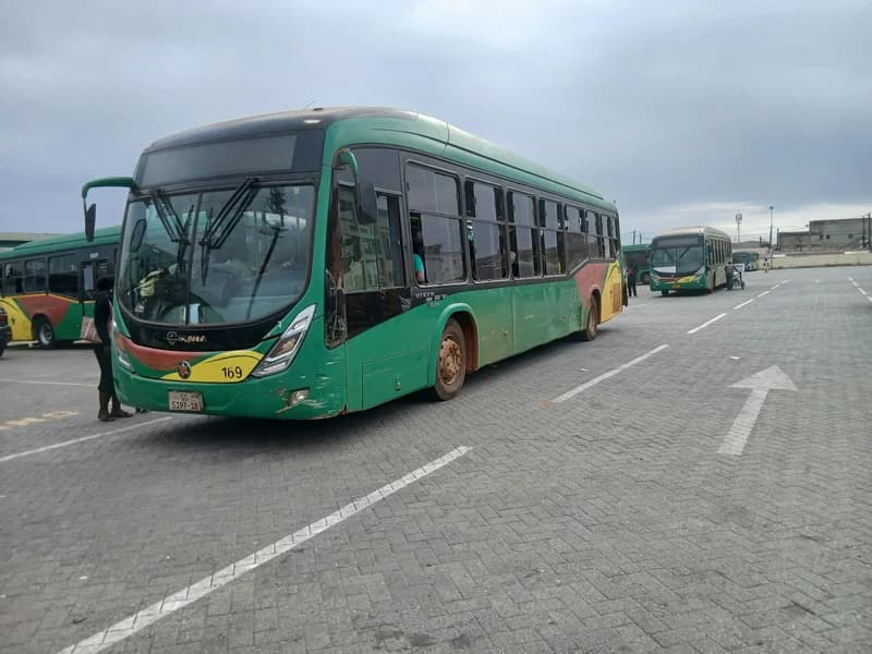 A green and yellow Aayalolo bus is parked at a paved terminal in Accra, with several other buses from the same fleet visible in the background. The main bus in the foreground is marked with the number 169 and shows a Ghanaian license plate under a cloudy sky.