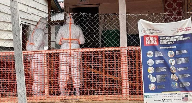 Two health workers in full white personal protective equipment (PPE) with orange markings stand behind a safety fence at a medical facility. Beside them, a large public health poster in French titled 'STOP EBOLA' outlines safety protocols and 'Bonnes pratiques' (good practices) for community protection.