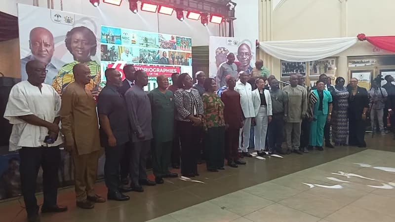 A large group of National Democratic Congress (NDC) members and officials stand together for a commemorative photo during a book launch event. The stage features a prominent backdrop with portraits of John Mahama and Naana Jane Opoku-Agyemang, alongside a collage titled 'Historiographies of the NDC'.