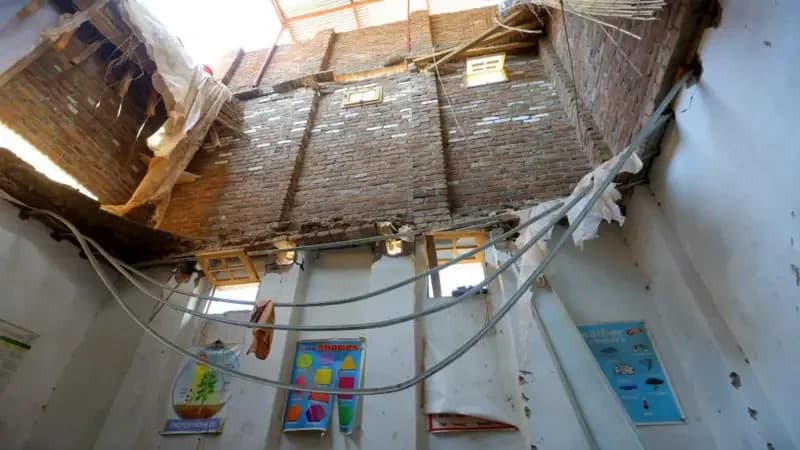 A photograph showing the interior of a classroom after a catastrophic roof collapse. The brick structure of the upper wall is exposed to the sky, and educational posters for children remain on the lower white walls amidst debris and hanging wires.