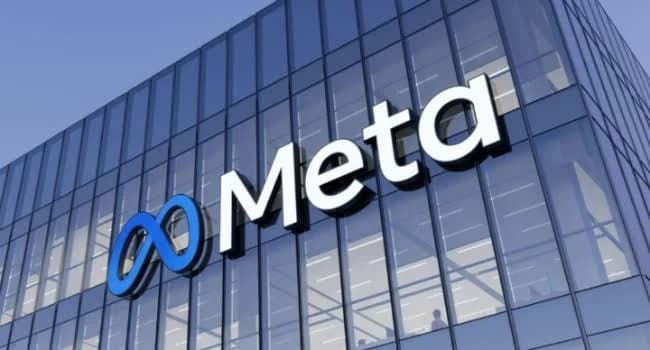 A photograph of a modern glass office building featuring the prominent 'Meta' wordmark and its blue infinity logo on the facade. The image represents the corporate identity of the technology company under a clear sky.