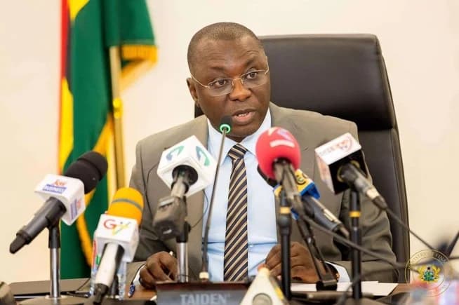 Dr. Mohammed Amin Adam is captured speaking during a press briefing, seated behind a row of microphones from various media houses including Joy Business and GTV. He is dressed in a grey suit and striped tie, with the Ghana national flag and the Ministry of Finance seal visible in the background.