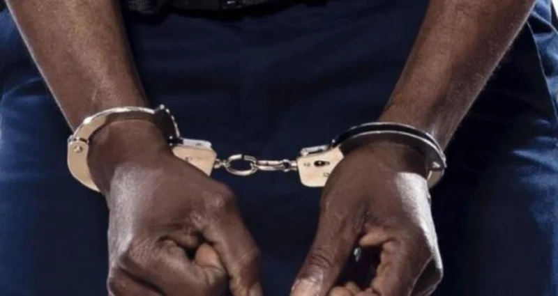 A close-up photograph showing a person's hands restrained in silver metallic handcuffs. The individual is wearing dark blue clothing, which is often associated with law enforcement or detention contexts.