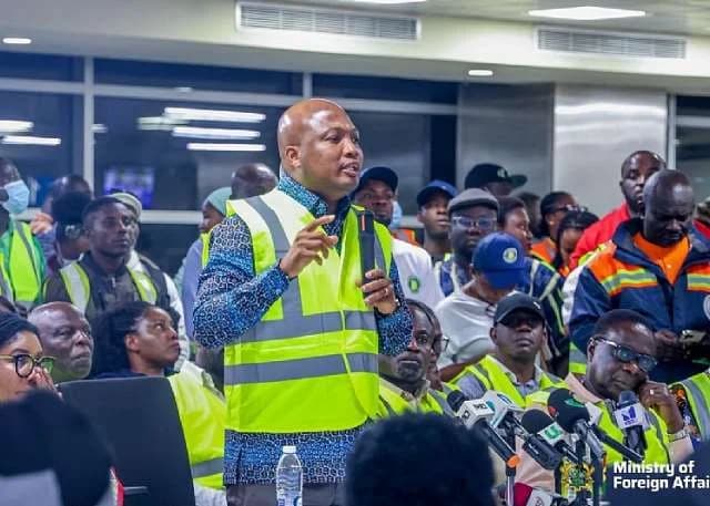 Samuel Okudzeto Ablakwa, MP for North Tongu, is captured speaking into a microphone while wearing a bright yellow high-visibility safety vest. He is surrounded by a crowd of people, many in similar safety gear, during what appears to be an official reception or briefing at an indoor facility like an airport.