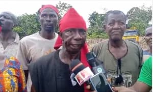 A group of farmers in Assin Kumasi are seen addressing the media while wearing red headbands and hats, which are traditional Ghanaian symbols of protest and grievance. The central figure is speaking into multiple microphones, with a yellow heavy-duty vehicle visible in the background near a wooded area.