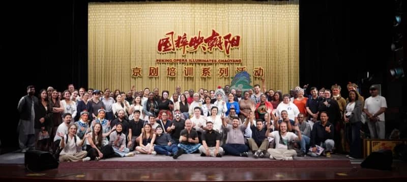 A large, diverse group of international journalists poses for a group photograph on a stage in front of a shimmering gold curtain. Several participants are seen wearing traditional Peking Opera costumes and face paint, indicating a cultural exchange and training session in the Chaoyang district of Beijing.