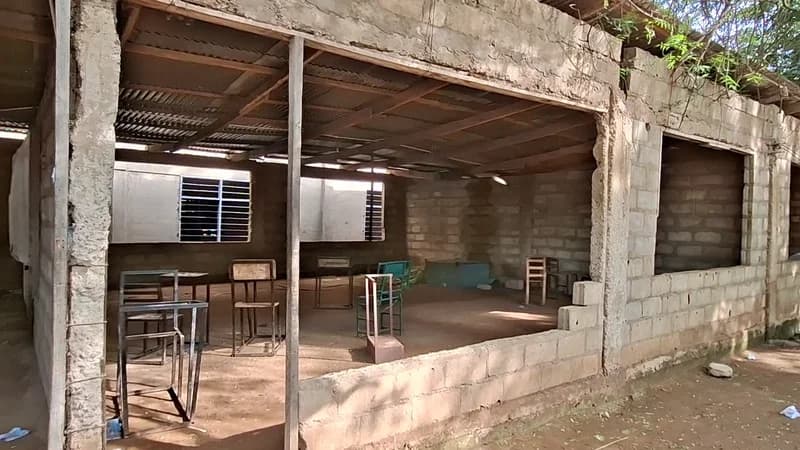 An unfinished and dilapidated classroom at Zamse Senior High School shows a severe shortage of desks and chairs. The structure features bare concrete block walls and a simple corrugated metal roof, with only a few pieces of worn furniture visible in the dusty interior.