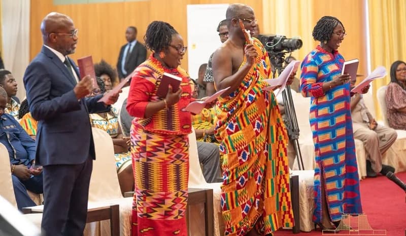 Four newly appointed Ghanaian envoys are shown standing during a formal swearing-in ceremony, holding holy books and documents as they recite their oaths. They are dressed in a mix of formal Western suits and colorful traditional Kente cloth, representing Ghana's cultural heritage in a diplomatic setting.