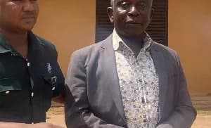 A photograph showing an older man in a grey suit jacket and a patterned shirt standing next to a man in a dark green uniform. The man in uniform appears to be escorting or detaining the man in the suit in front of a tan building.