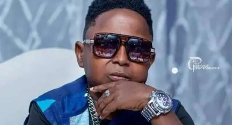 A close-up portrait of Ghanaian Kumawood actor Wayoosi wearing dark sunglasses and a blue jacket. He is pictured with his hand to his chin, showcasing a large silver wristwatch and a chain necklace.