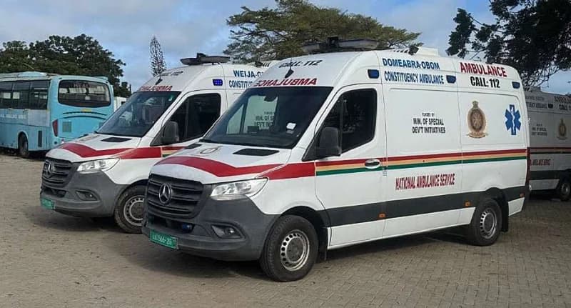 Two white Mercedes-Benz Sprinter ambulances belonging to the National Ambulance Service of Ghana are parked in an outdoor lot. The vehicles are labeled for the Domeabra-Obom and Weija-Gbawe constituencies, showcasing the fleet used for emergency medical services in the region.
