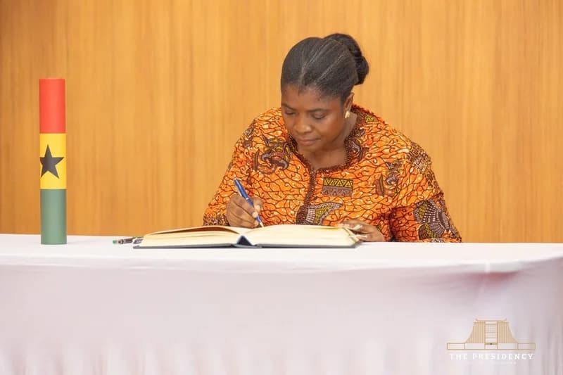 Emelia Agbenyo is seen signing the official oath book during her swearing-in ceremony as a Deputy Electoral Commission (EC) Chair. She is dressed in a vibrant orange patterned traditional outfit, seated at a table covered with a white cloth, with a Ghana flag-themed cylinder placed beside her.