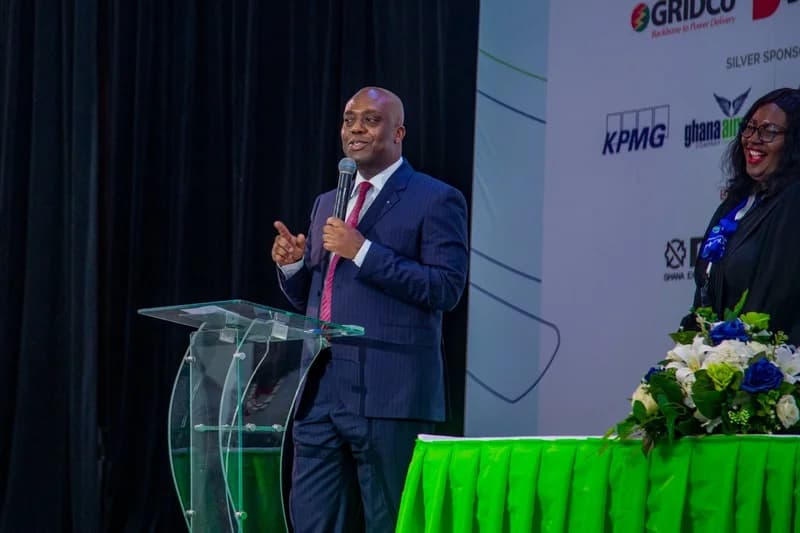 Deputy Finance Minister Dr. Stephen Amoah is seen speaking at a formal business event, holding a microphone while standing behind a glass lectern. He is dressed in a navy pinstripe suit and red tie, with corporate sponsor logos for organizations like GRIDCo and KPMG visible on the backdrop behind him.